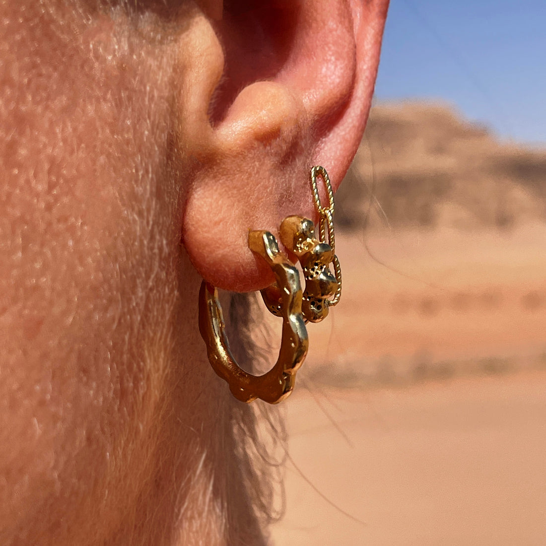 Photograph of and ear with three earrings, a gold coral inspired hoop earrings, a smaller gold hoop with texure and a small chain stud earring all in gold, with the desert in the back ground.