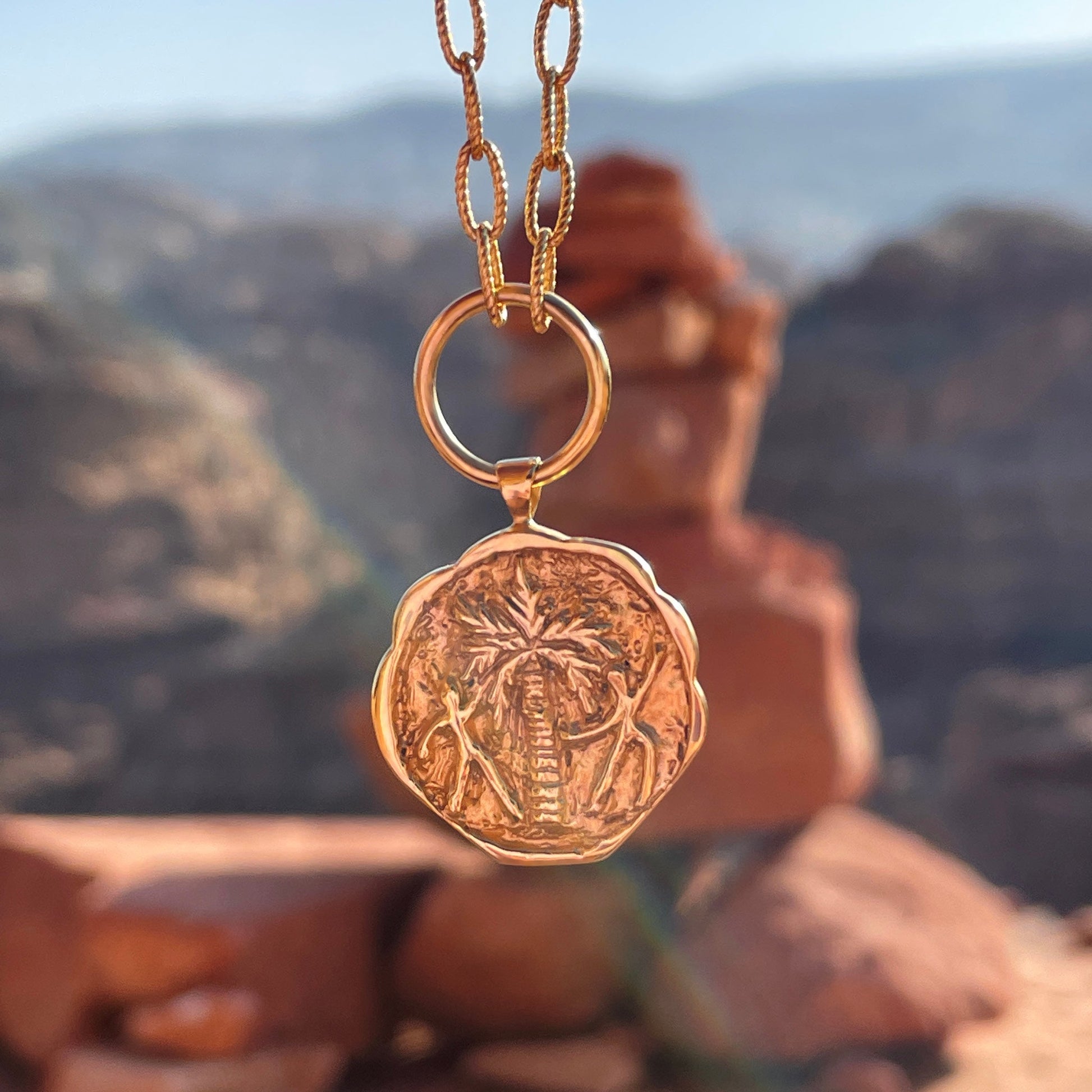 Gold coin pendant necklace engraved with a palm tree and two people dancing around it. In the background, a desert landscape.