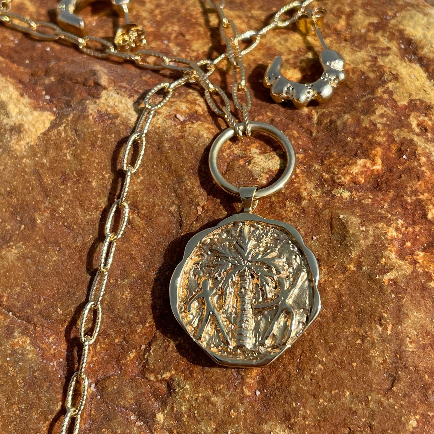 Gold coin pendant necklace engraved with a palm tree and two people dancing around it, laying on a a rock. Gold textured huggie earrings in the background.