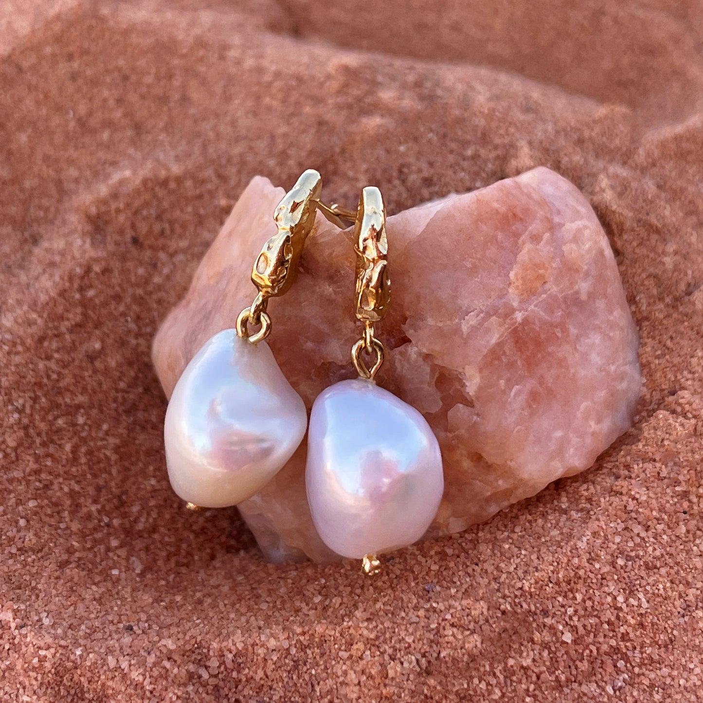 Gold textured earrings with large baroque pearls hanging below, resting on a piece of rose quartz and terracotta sand.