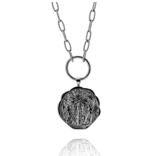 Silver coin pendant necklace engraved with a palm tree and two people dancing around it on a white background.