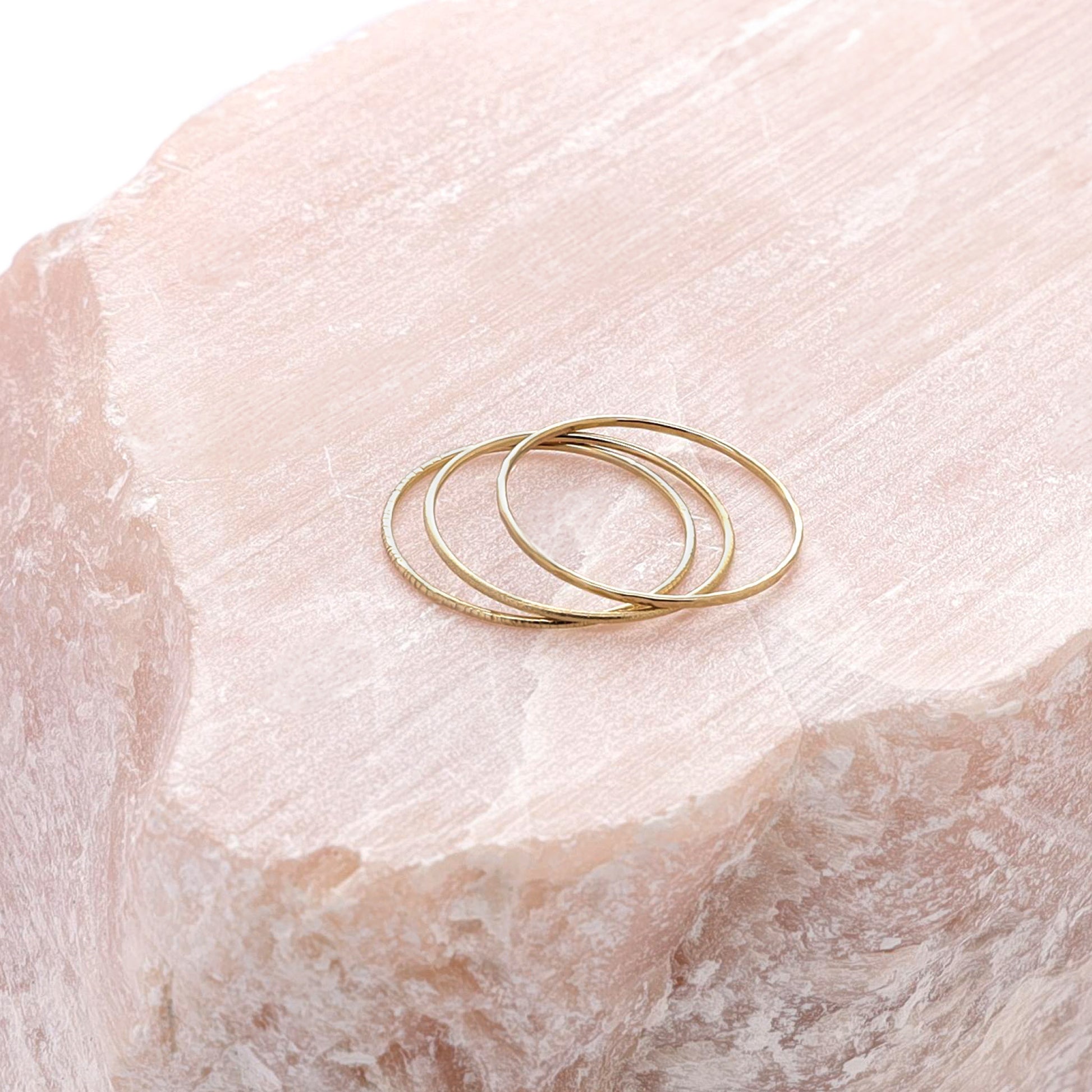 Three slim gold stacking rings, each with a different texture, displayed on a large piece of rose quartz.