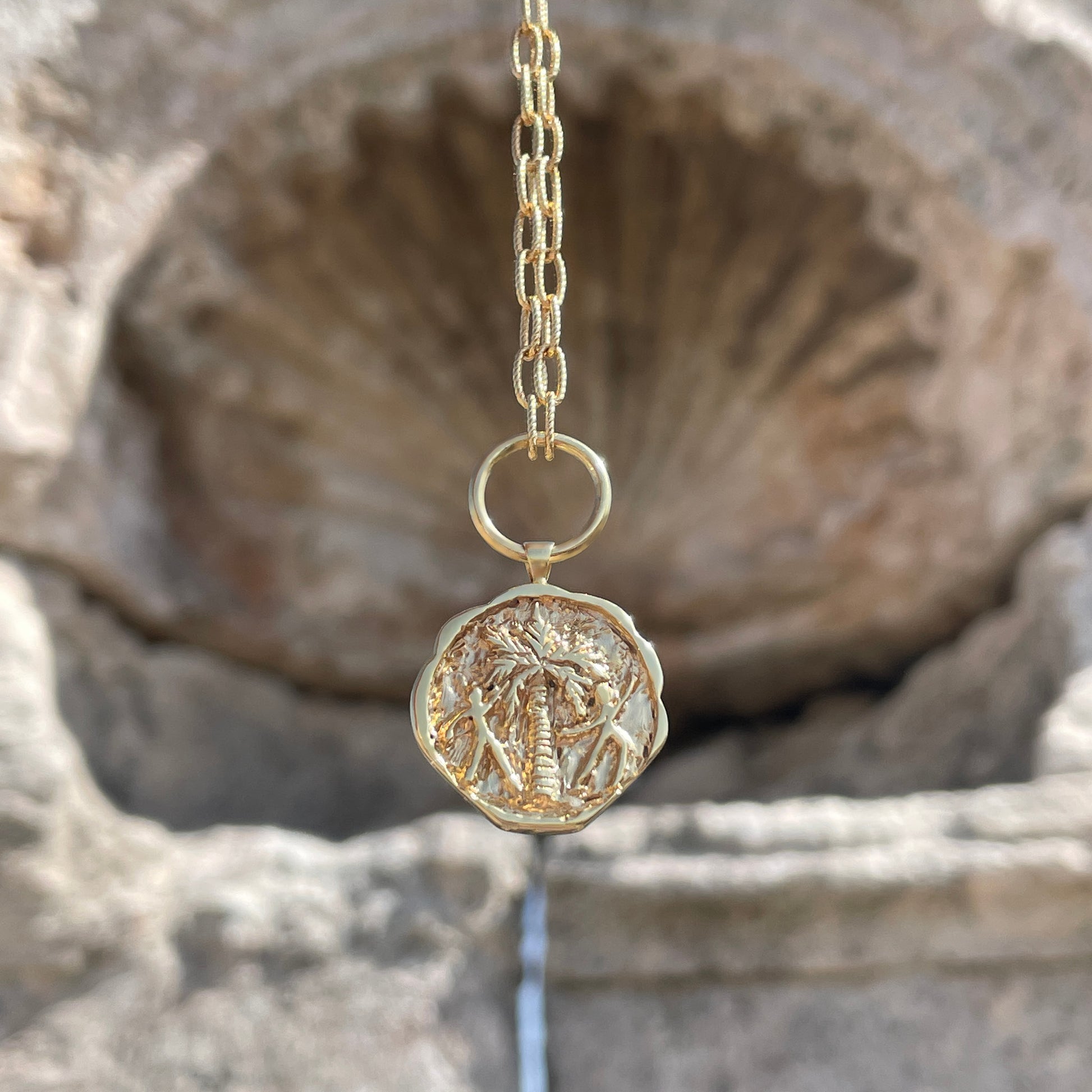 Gold coin pendant necklace engraved with a palm tree and two people dancing around it.